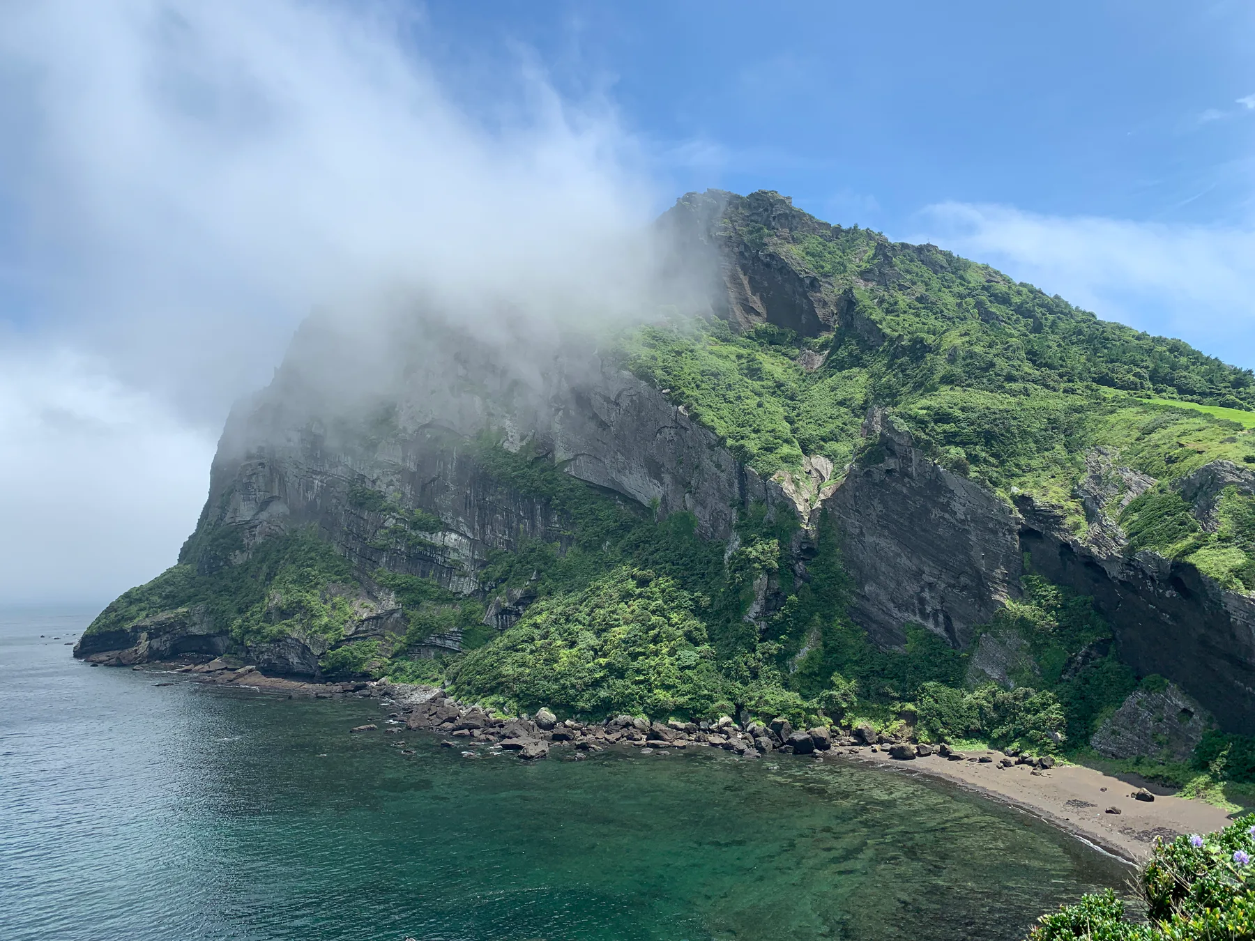 A volcanic coastal landscape in Jeju with clouds over green cliffs.