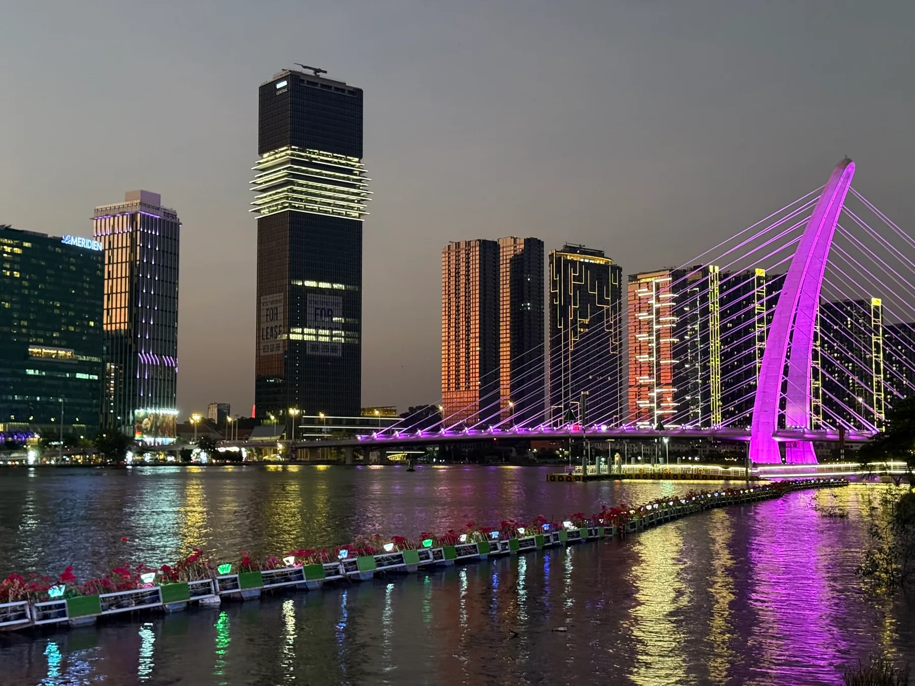 Ho Chi Minh City buildings and a lit bridge reflected in the river after dark.